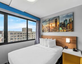Queen bed beside large city-view window with skyline artwork and bright accents at Days Inn Chinatown NYC.