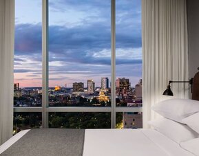 Bedroom with floor-to-ceiling windows showcasing evening skyline views at Moxy Boston Downtown.