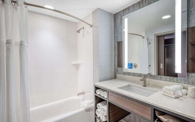 Modern bathroom with illuminated mirror, bathtub shower combo, and marble-style vanity at Hilton Garden Inn Boston Logan Airport.