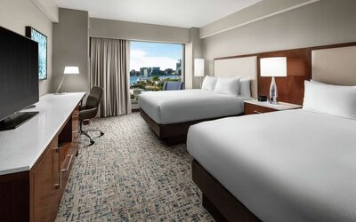 Room with two beds, workspace desk, and waterfront city view through large window at Hilton Boston Logan Airport.