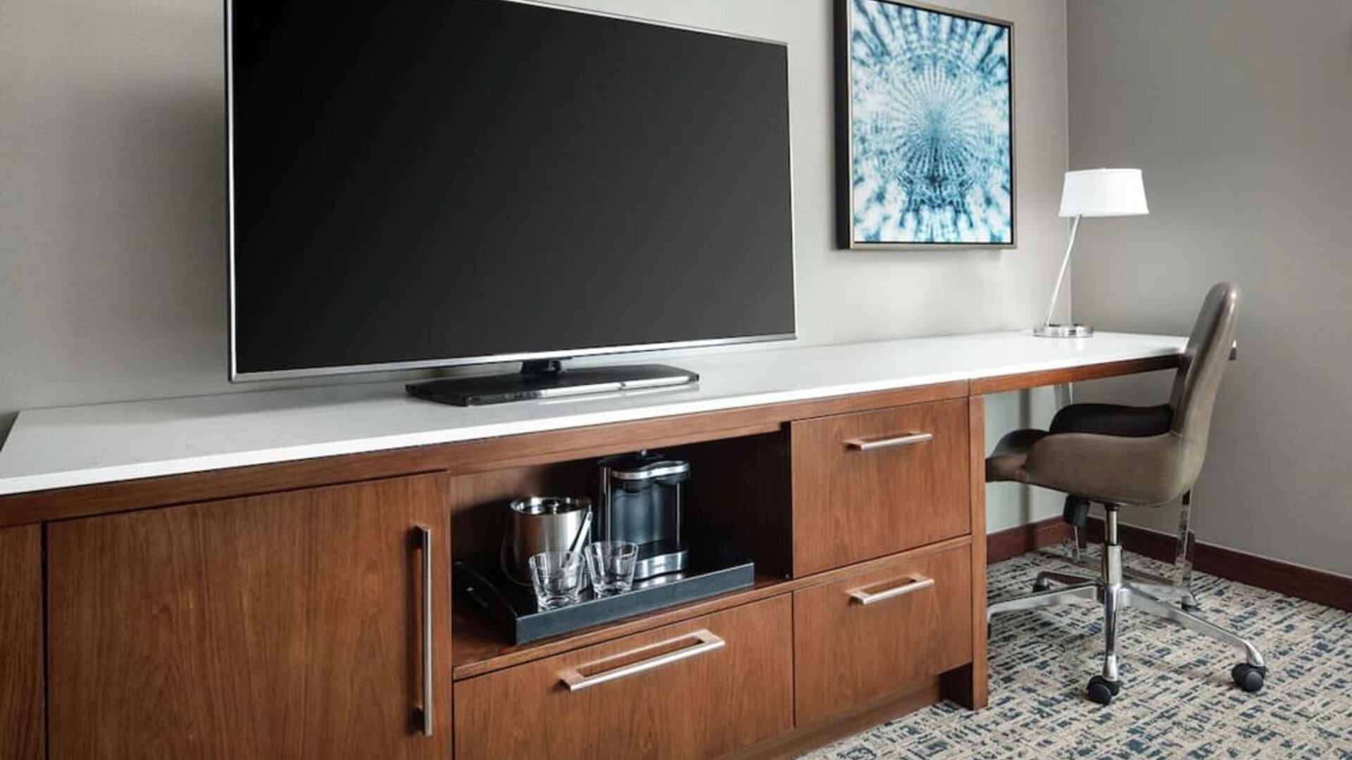Workspace with desk chair, large TV, coffee station, and wooden cabinetry at Hilton Boston Logan Airport.