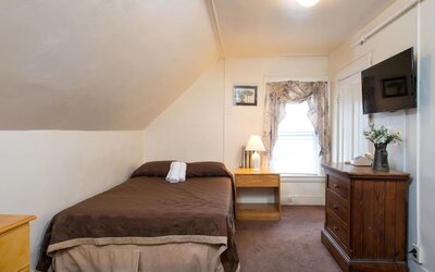 Small attic-style guest room with double bed, wooden dresser, and wall-mounted TV at The Farrington Inn.