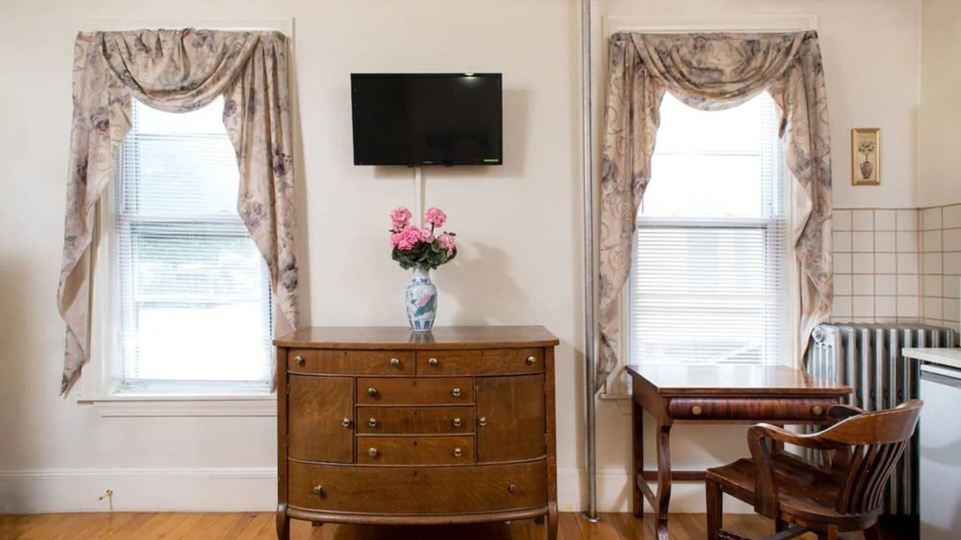Seating and workspace area with wooden desk, dresser, floral curtains, and mounted TV at The Farrington Inn.