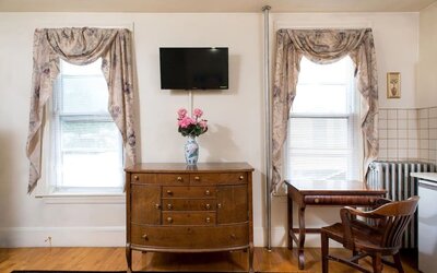 Seating and workspace area with wooden desk, dresser, floral curtains, and mounted TV at The Farrington Inn.