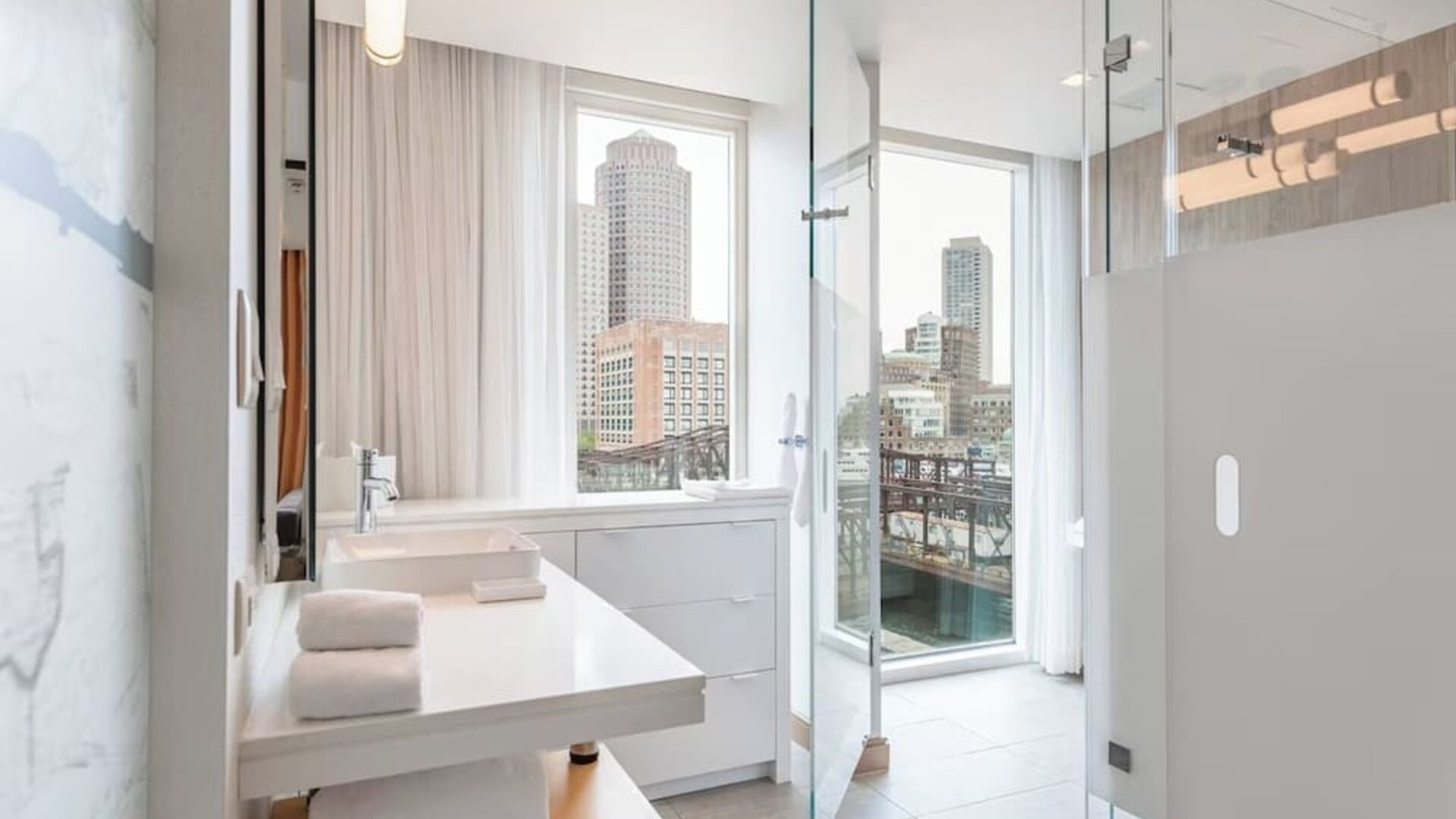 Sleek modern bathroom with floor-to-ceiling windows, glass shower enclosure, and stunning Boston waterfront views at The Envoy Hotel, Autograph Collection.