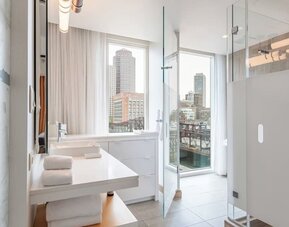 Sleek modern bathroom with floor-to-ceiling windows, glass shower enclosure, and stunning Boston waterfront views at The Envoy Hotel, Autograph Collection.
