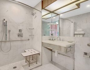 Accessible guest bathroom at Wyndham Garden Brooklyn Sunset Park featuring a roll-in shower, safety grab bars, shower seating, marble finishes, and a spacious vanity designed for comfort and accessibility.