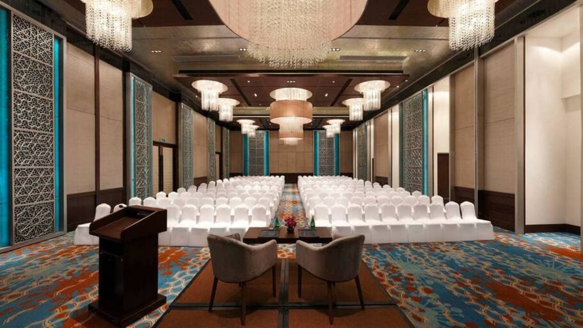 Long presentation and banquet hall with ornate chandeliers, a podium and sitting area for two panelists at the front, at the Hilton Jaipur, India.