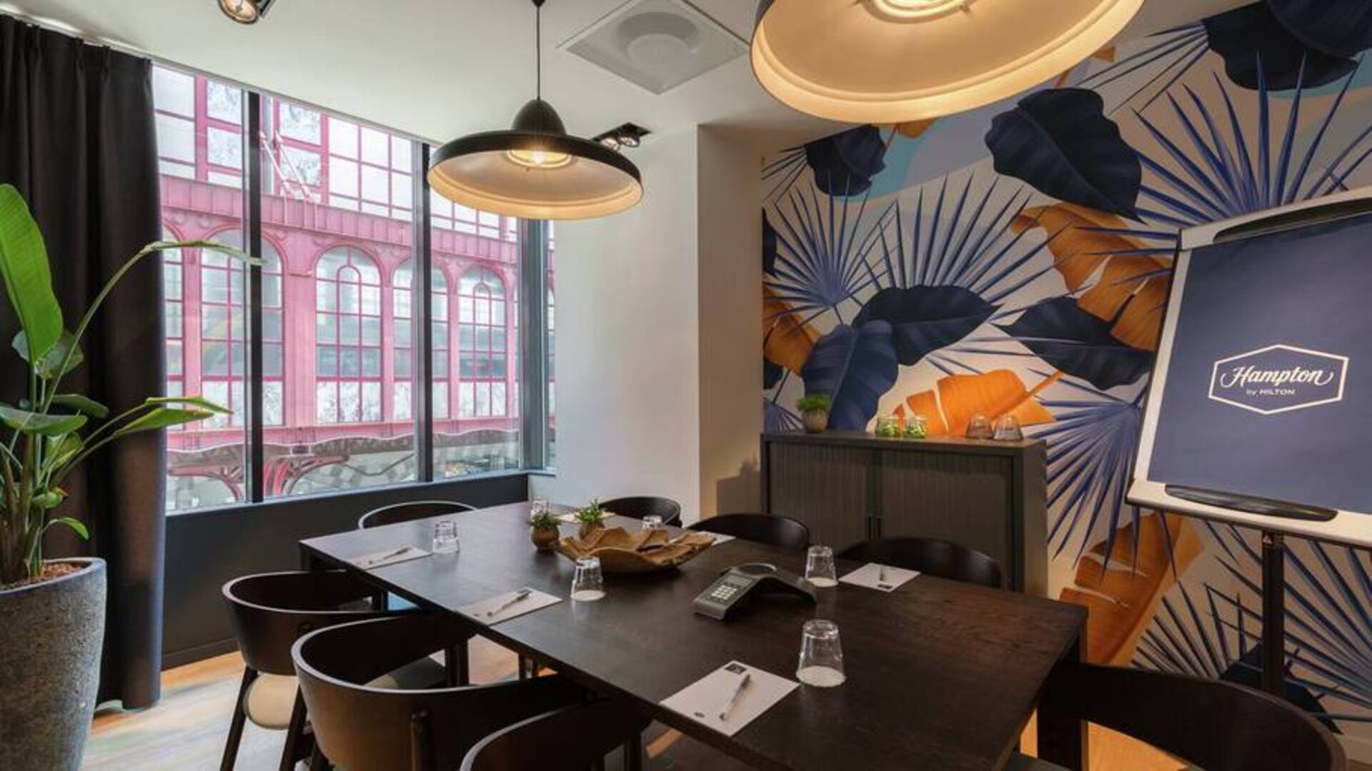 Floral artistic wall pattern and large window view of Antwerp complements a dark brown wooden table and place settings for 8 people to meet and do business in.