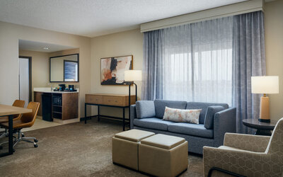 Spacious suite living area with comfortable sofa seating, work space, mini refreshment station, and large windows providing natural light at Embassy Suites By Hilton Charleston Airport Convention Center.