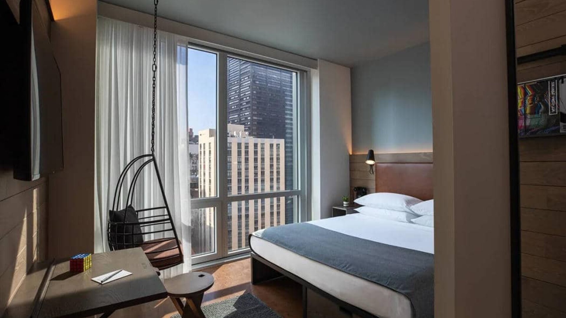 Moxy NYC Downtown double bed guest room, with large television, floor-to-ceiling window, and desk for working.