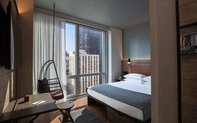 Moxy NYC Downtown double bed guest room, with large television, floor-to-ceiling window, and desk for working.