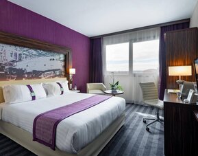 Guest room with a double bed, purple accents, airplane artwork, large window, lounge chair, work desk, and TV at Holiday Inn Toulouse Airport.
