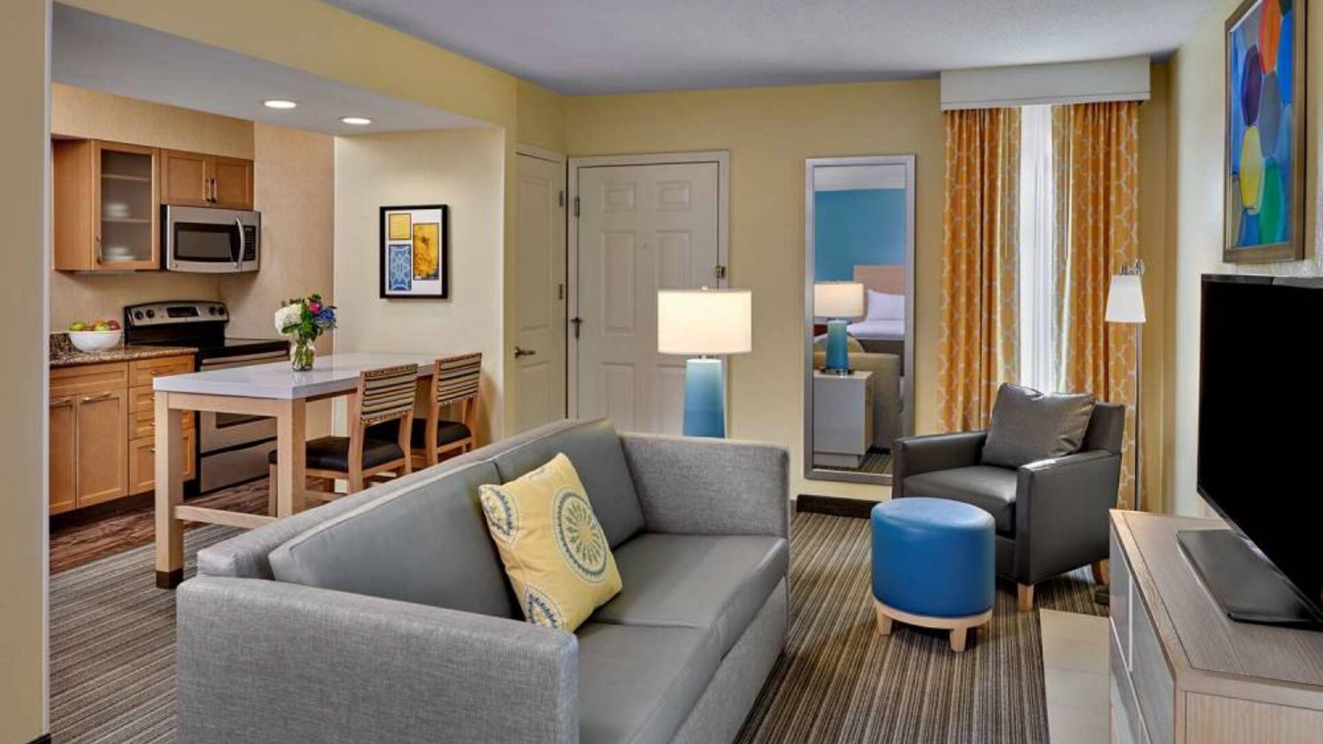Sonesta ES Suites Cincinnati - Blue Ash guest room lounge, with armchair, sofa, and television, plus nearby kitchenette.