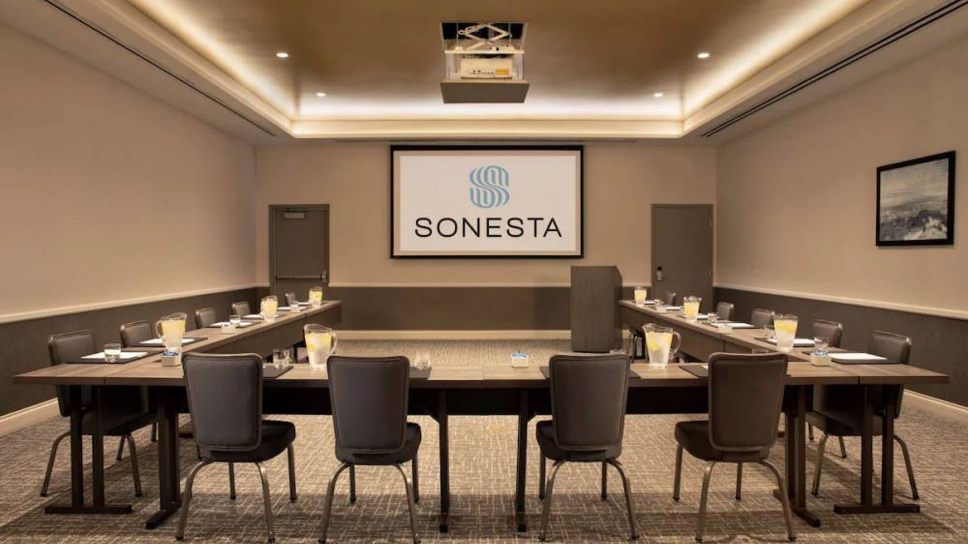 Hotel meeting room with long tables arranged in a U-shape, facing a large projector screen.