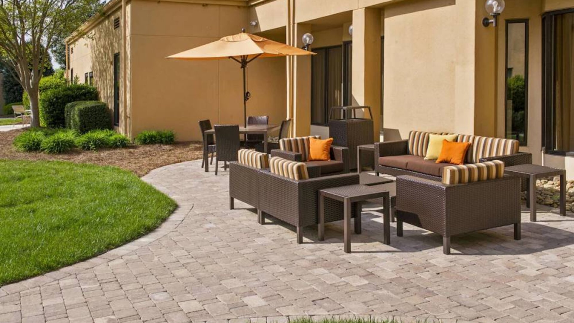 The patio at Sonesta Select Charlotte University Research Park includes sofa and armchair seating, plus coffee tables, near to pleasant greenery.