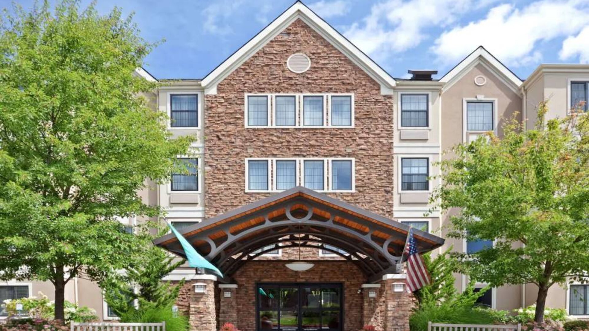 The hotel’s exterior has a covered entrance, wooden benches, and pleasant greenery including flowers and trees.