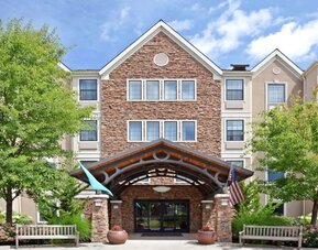 The hotel’s exterior has a covered entrance, wooden benches, and pleasant greenery including flowers and trees.
