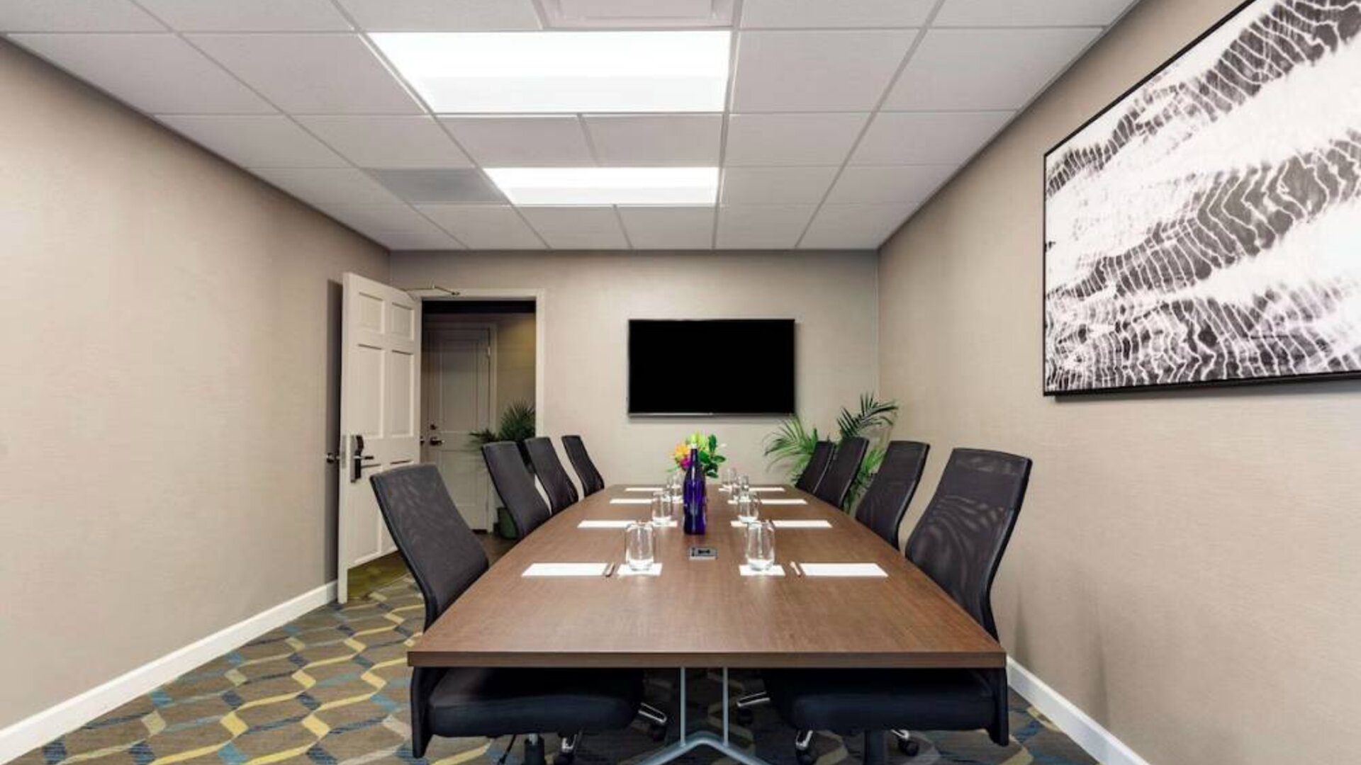 Hotel meeting room, featuring long wooden table, eight swivel chairs, and a large, widescreen TV on the wall.