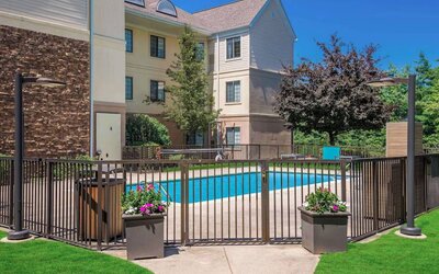 Sonesta ES Suites Andover Boston’s outdoor pool is ringed by a fence, with a backdrop of the hotel and numerous trees.