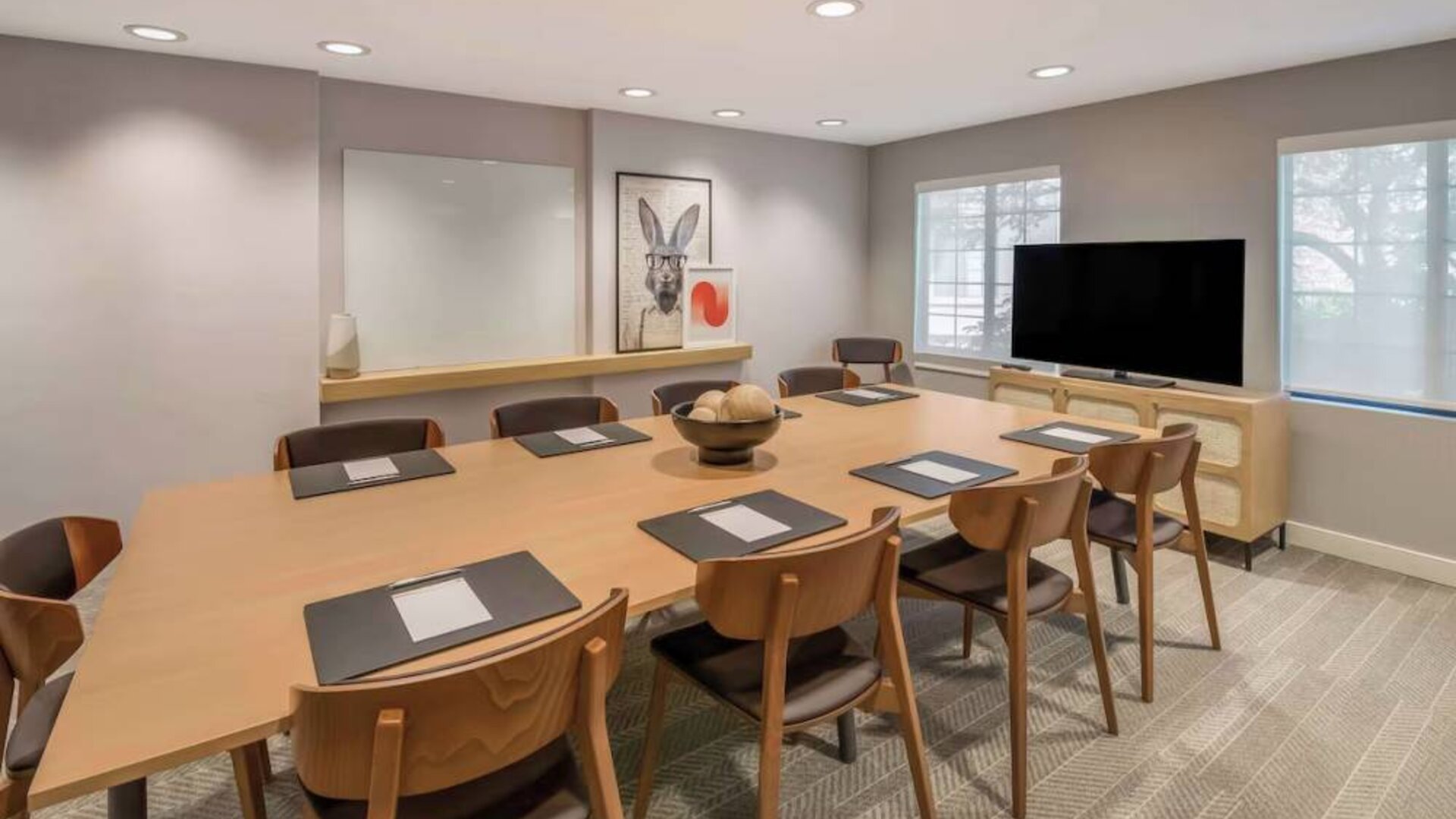 Hotel meeting room, with long wooden table and seating for nine, art and a whiteboard on the wall, and a large television. 
