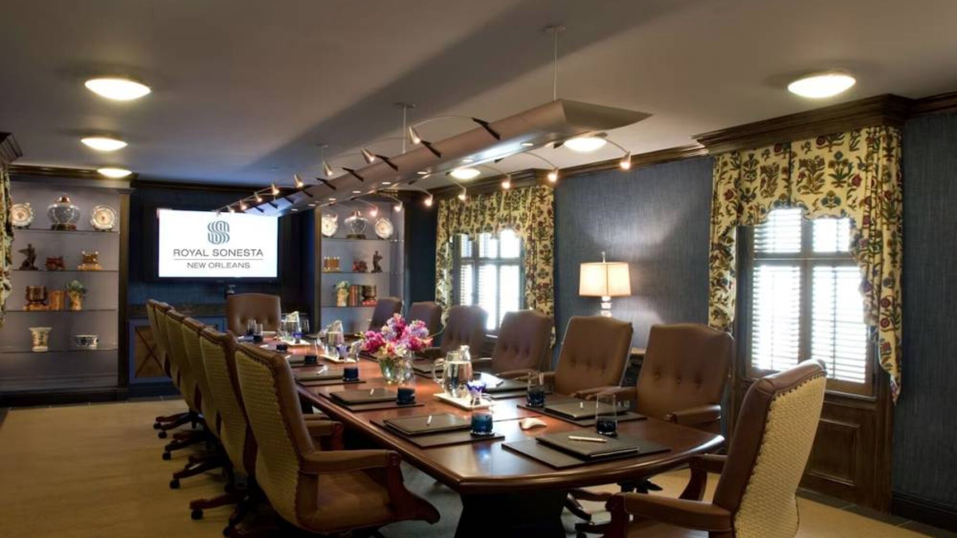 Meeting room in Royal Sonesta New Orleans, furnished with long wooden table, 14 swivel chairs, a widescreen TV, and two windows.