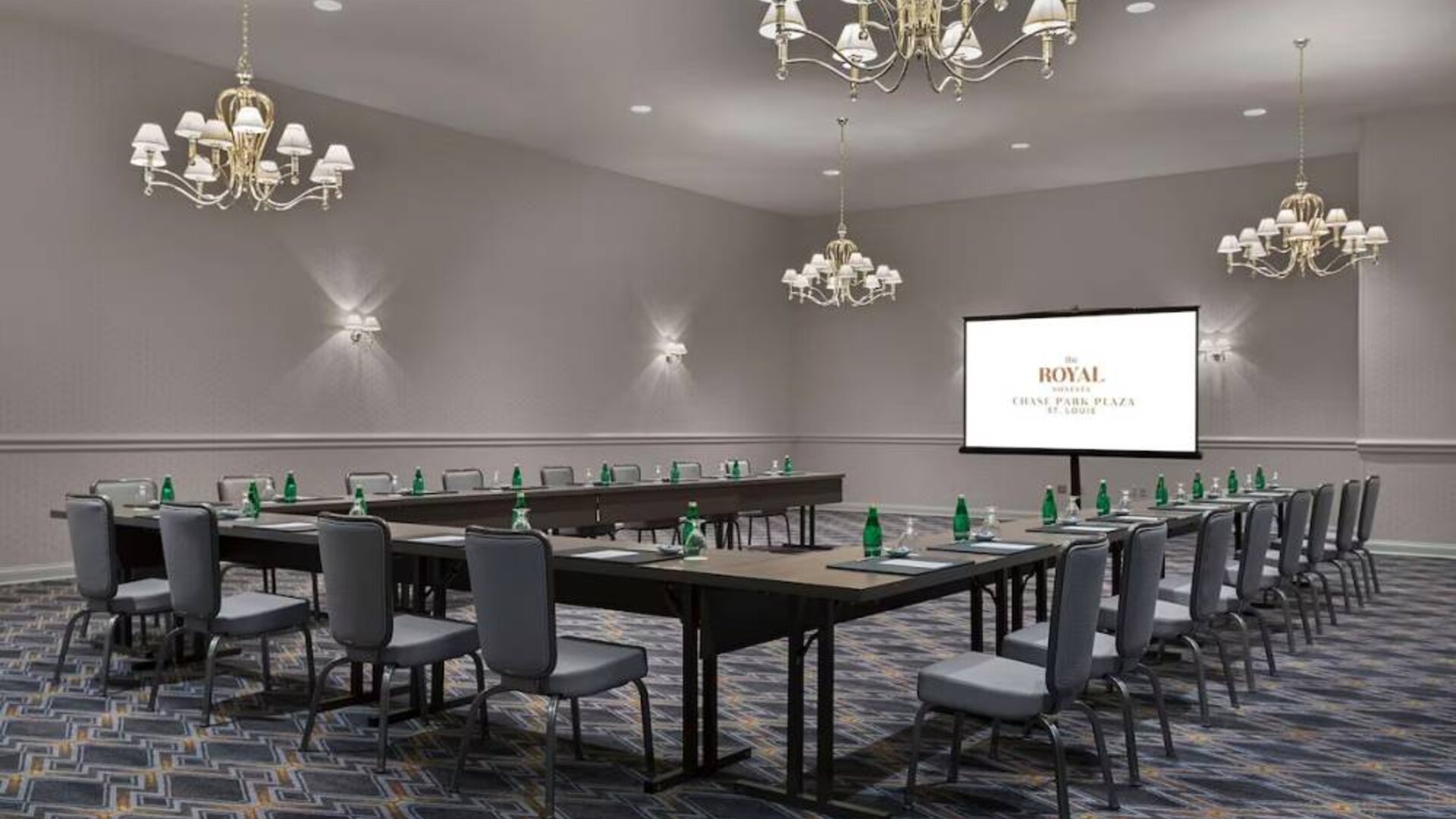 Meeting room in The Chase Park Plaza Royal Sonesta St. Louis, featuring long tables arranged in a U-shape, quartet of chandeliers, seating for over a dozen, and a projector screen.