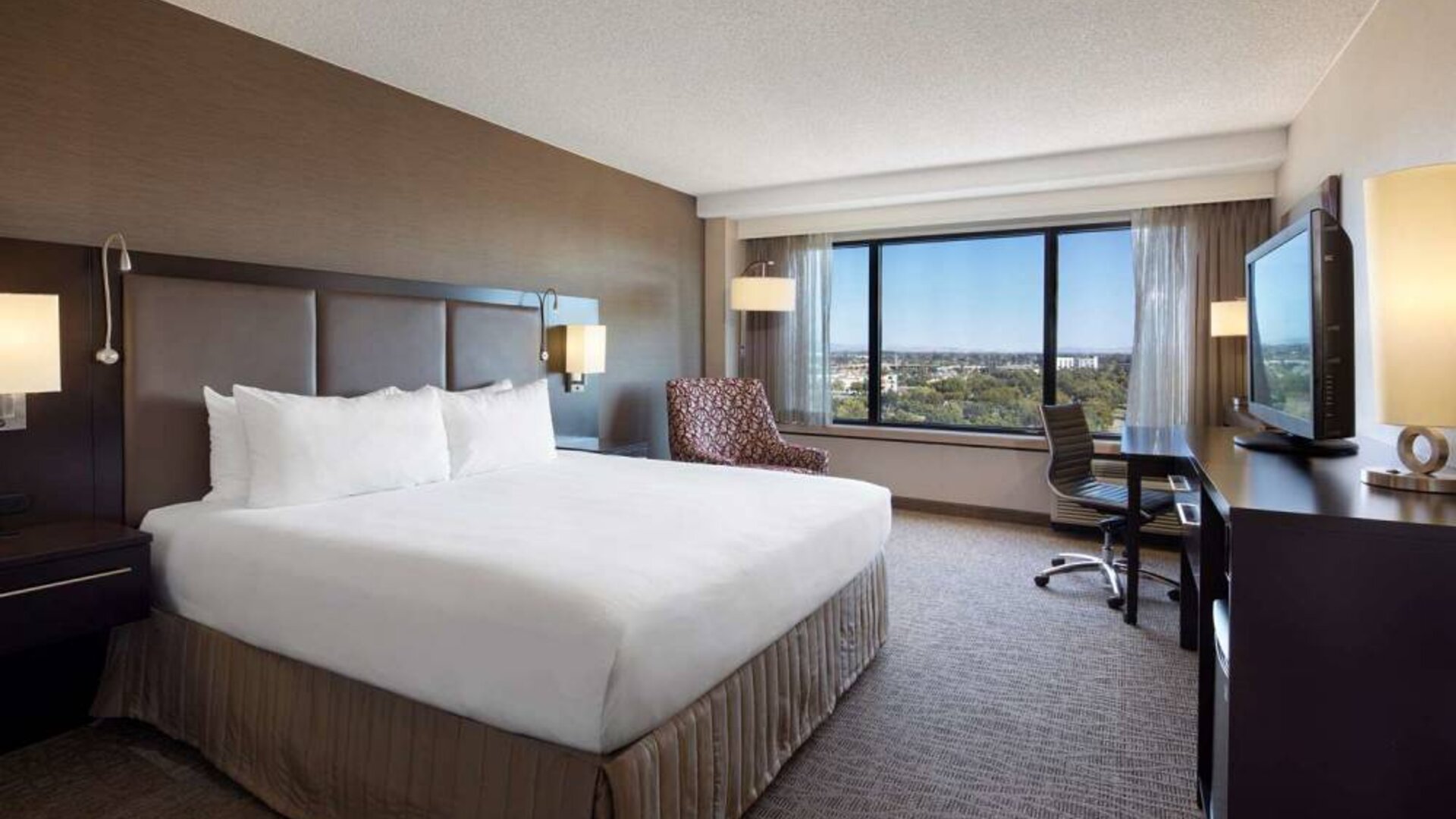 Double bed guest room Sonesta San Jose, with workspace desk and chair, armchair, TV, and window.