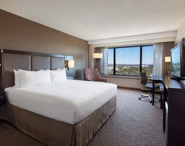 Double bed guest room Sonesta San Jose, with workspace desk and chair, armchair, TV, and window.