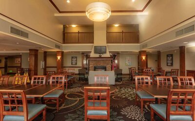 The hotel breakfast area has a high ceiling, wide range of table sizes, carpeted floor, and a wall-mounted TV.