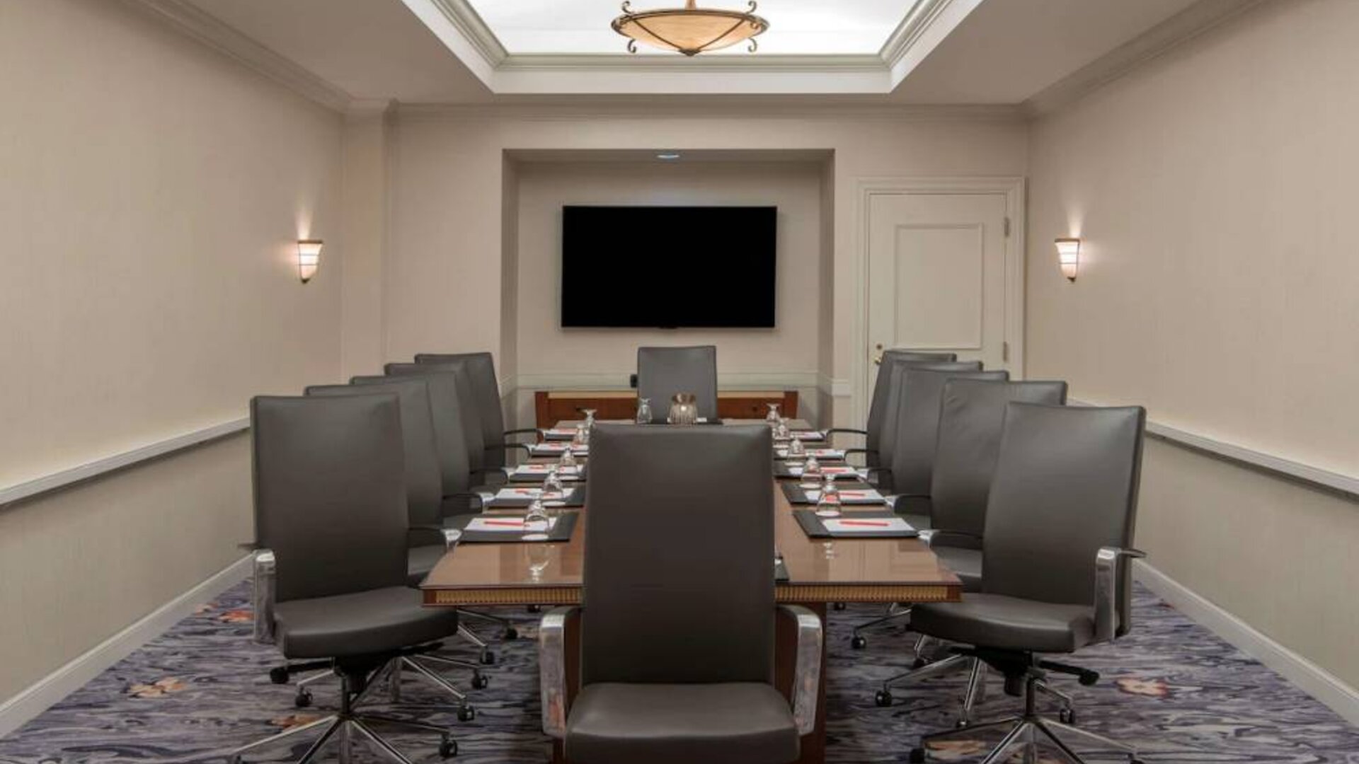 Sonesta Charlotte Executive Park meeting room, furnished with long table, a dozen leather swivel chairs, and a large, wall-mounted TV.