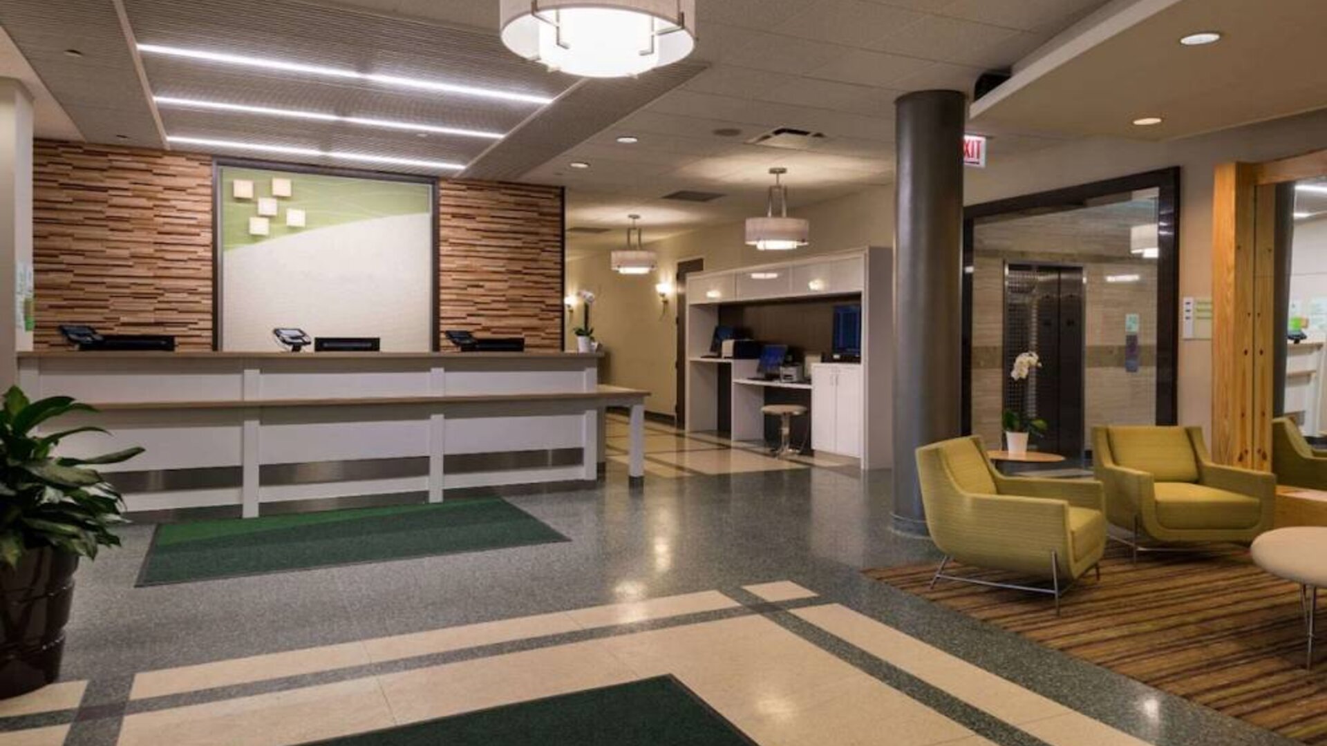 The hotel lobby has yellow armchairs, potted plants, and a business center workstation, in addition to the reception desk.