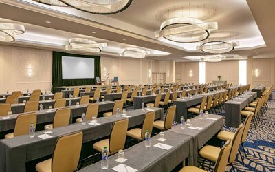 Hotel meeting room, furnished with tables and seating for dozens of attendees, arranged in a classroom style facing a large projector screen.