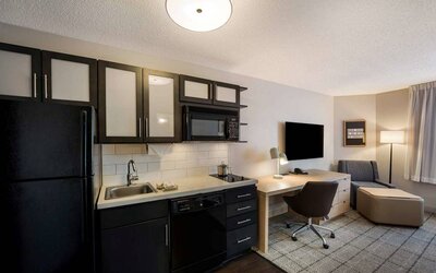 Sonesta Simply Suites Jersey City guest room kitchen, with microwave, oven, hob, and fridge-freezer, plus nearby chair and TV.