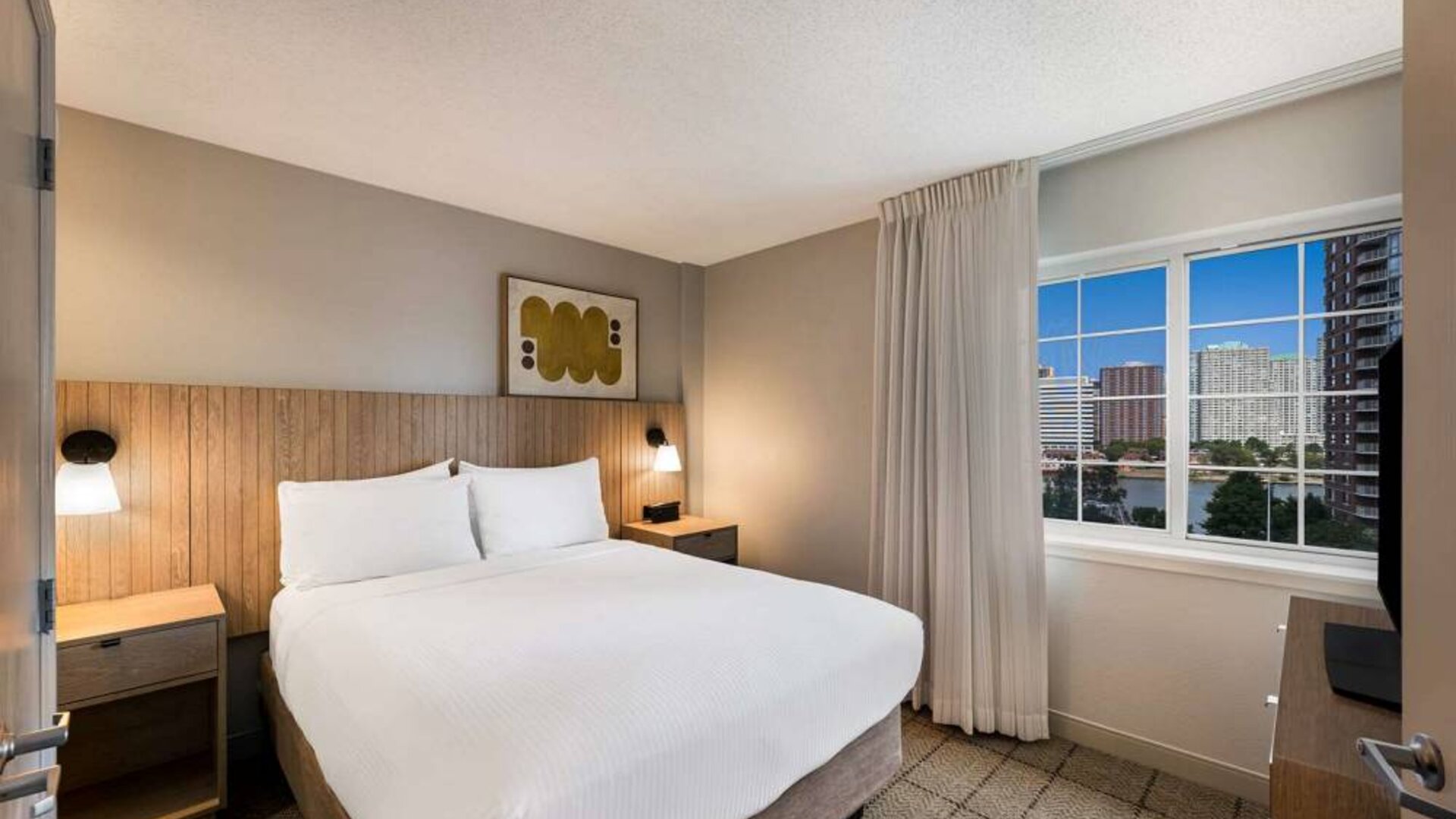 Double bed guest room in Sonesta Simply Suites Jersey City, including art on the wall, TV, and window.