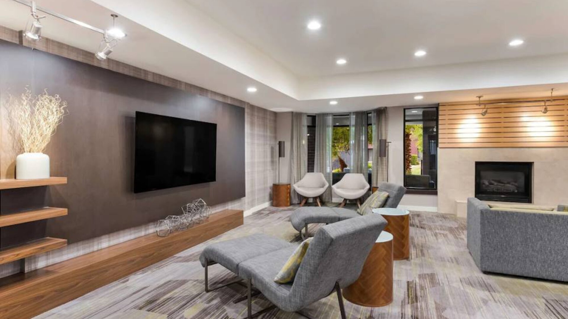 The hotel lobby includes a large, wall-mounted TV faced by a pair of reclined seats, with a fireplace and sofa nearby.