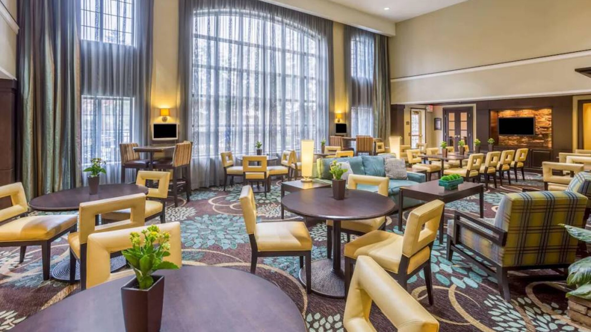 The hotel’s breakfast area has a carpeted floor, range of table sizes, seating from normal chairs to tall stools and armchairs, high ceilings and large windows, plus multiple TVs.