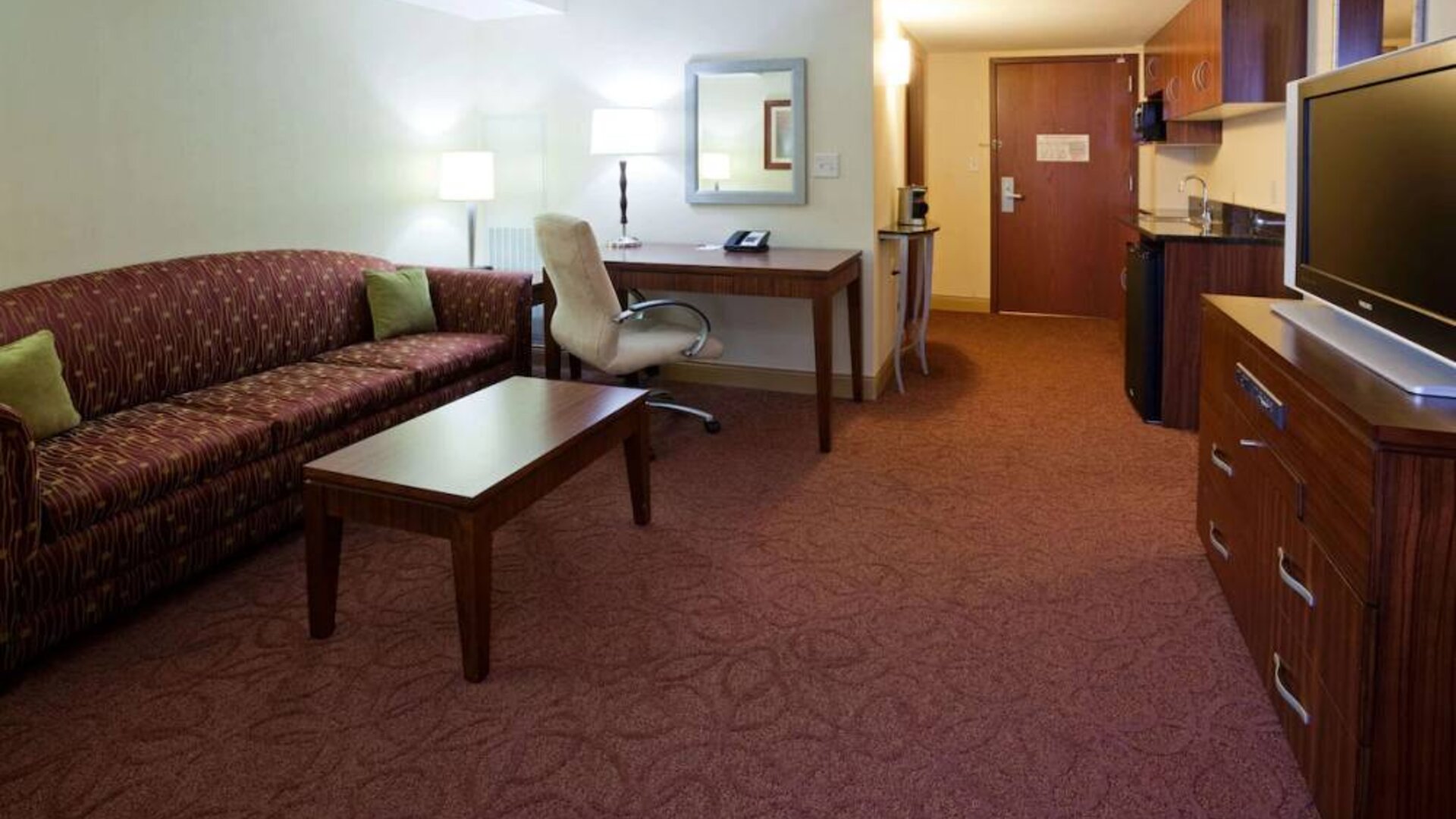 Sonesta Milwaukee West Wauwatosa guest room workspace with desk, chair, lamp, and telephone, with TV and sofa nearby.