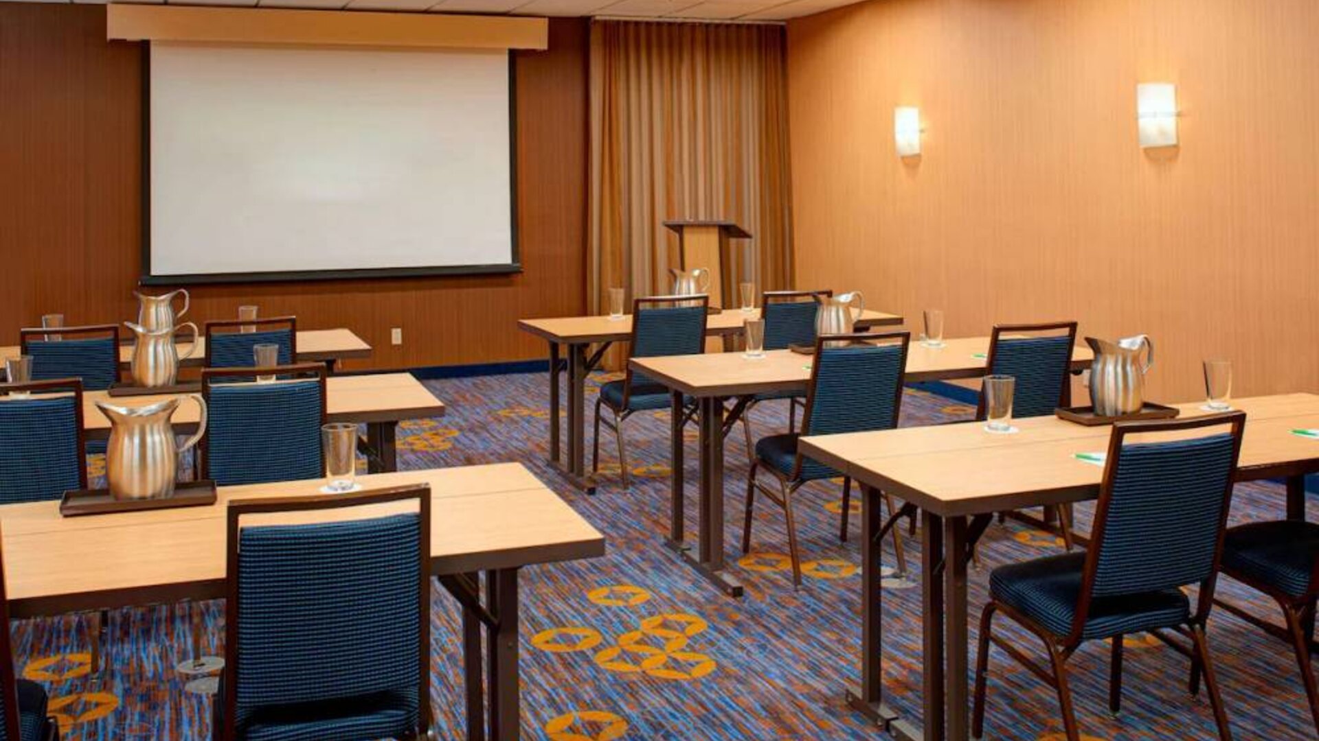 Hotel meeting room with seating for a dozen attendees and tables arranged in a classroom format facing a projector screen.