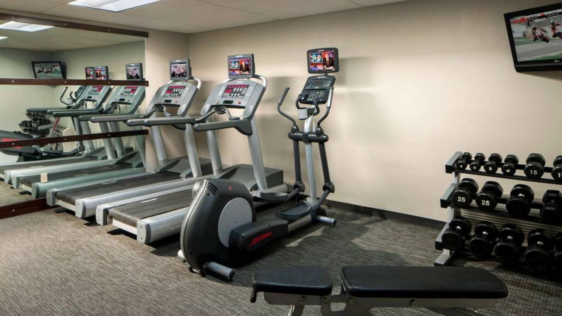 The fitness center at Sonesta Select Boca Raton comes with a wall-mounted TV, a variety of exercise machines, and rows of free weights.