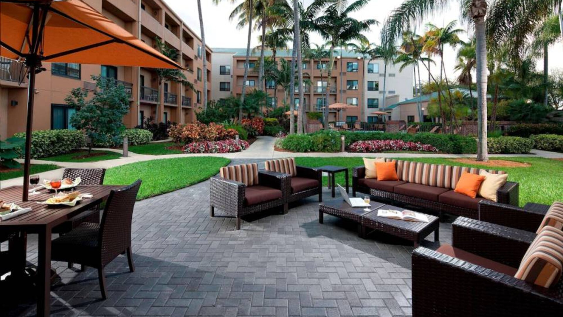 Patio area furnished with sofa and armchair seating and shaded table and chairs, close to pleasant greenery at Sonesta Select Miami Lakes.