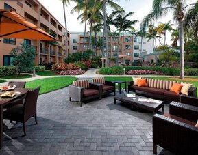 Patio area furnished with sofa and armchair seating and shaded table and chairs, close to pleasant greenery at Sonesta Select Miami Lakes.