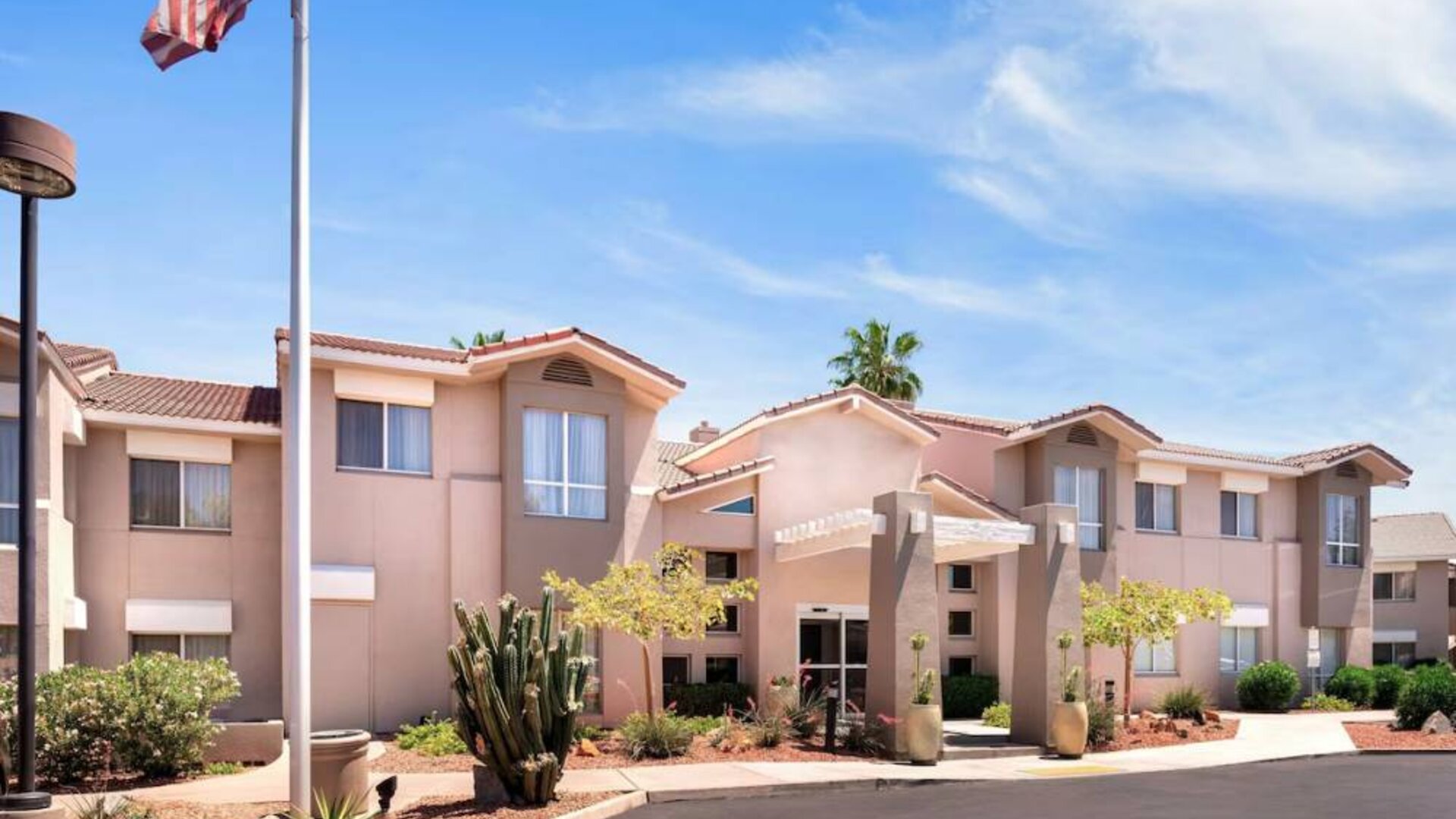 The hotel’s exterior has a fluttering USA flag flying from a flagpole and greenery including bushes, small trees, and a cactus.