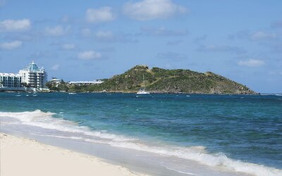 Waves coming on shore with view of resort buildings and mountainous island in distance. 