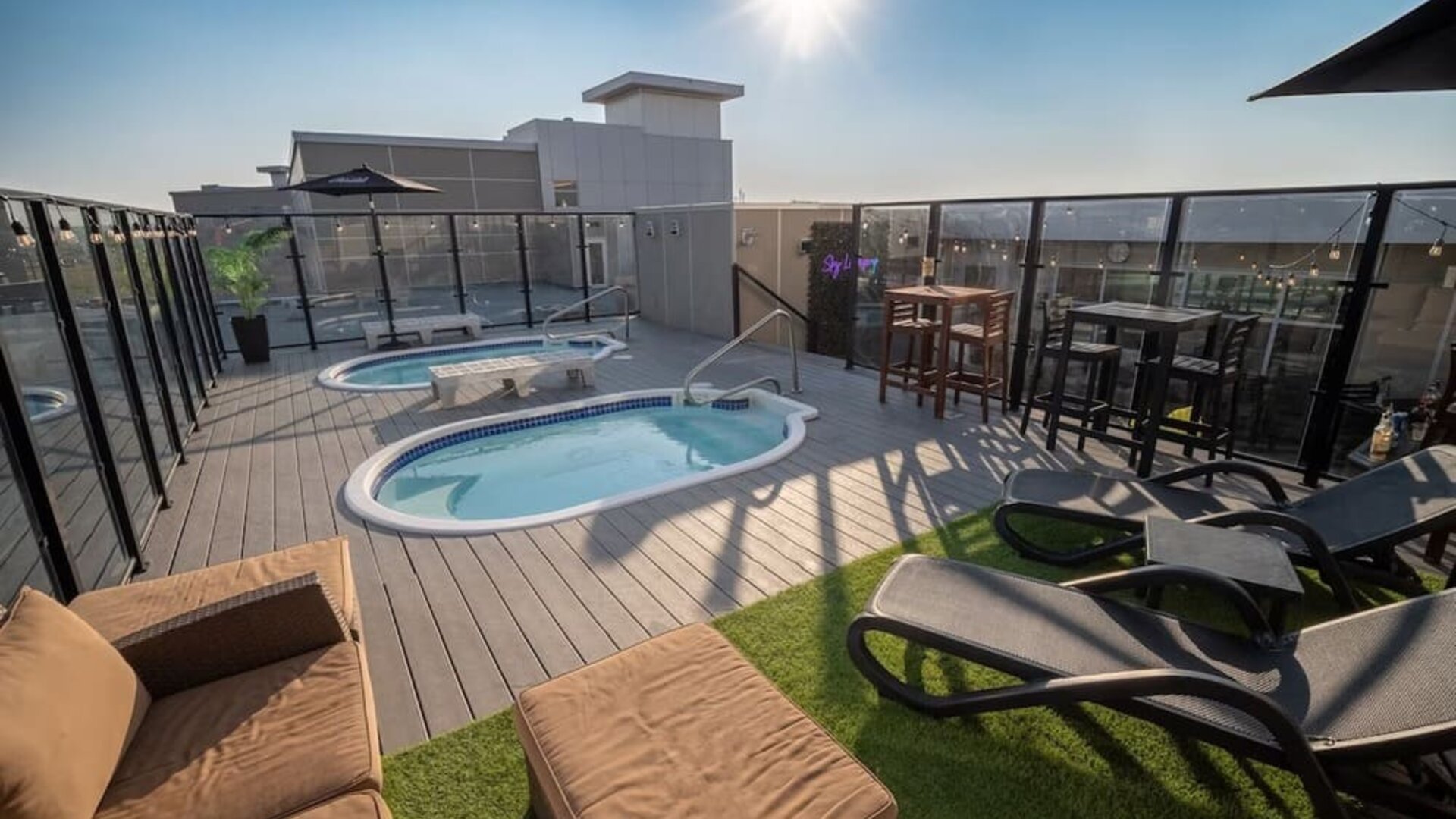 Rooftop deck with two hot tubs, lounge seating, high-top tables, and glass safety railing in daylight. Hotel Clique Calgary Airport.