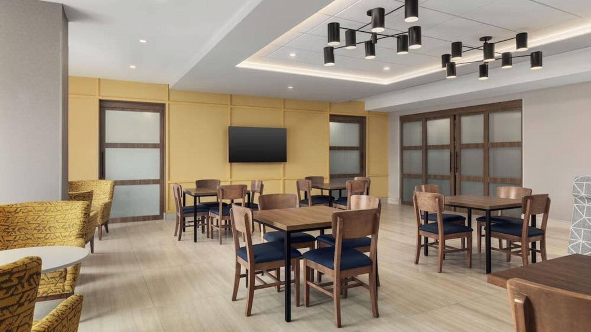 Bright breakfast dining area with wooden tables, cushioned chairs, and modern ceiling lighting at Hampton Inn By Hilton Montreal East Anjou.