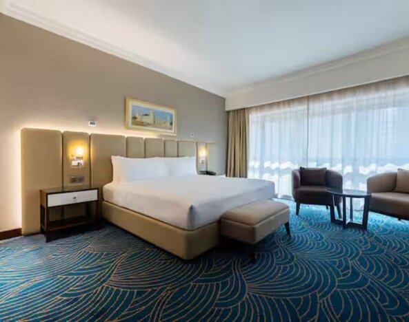Spacious king room with floor-to-ceiling windows, lounge seating, patterned carpet, and natural daylight.