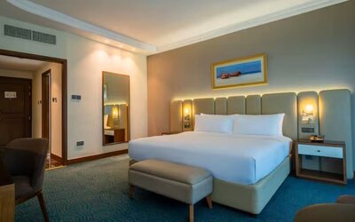 Bright guest room featuring a king bed, upholstered headboard, bedside lighting, seating area, and workspace.