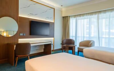 Contemporary guest room with two twin beds, large window, seating area, wall-mounted television, and workspace.