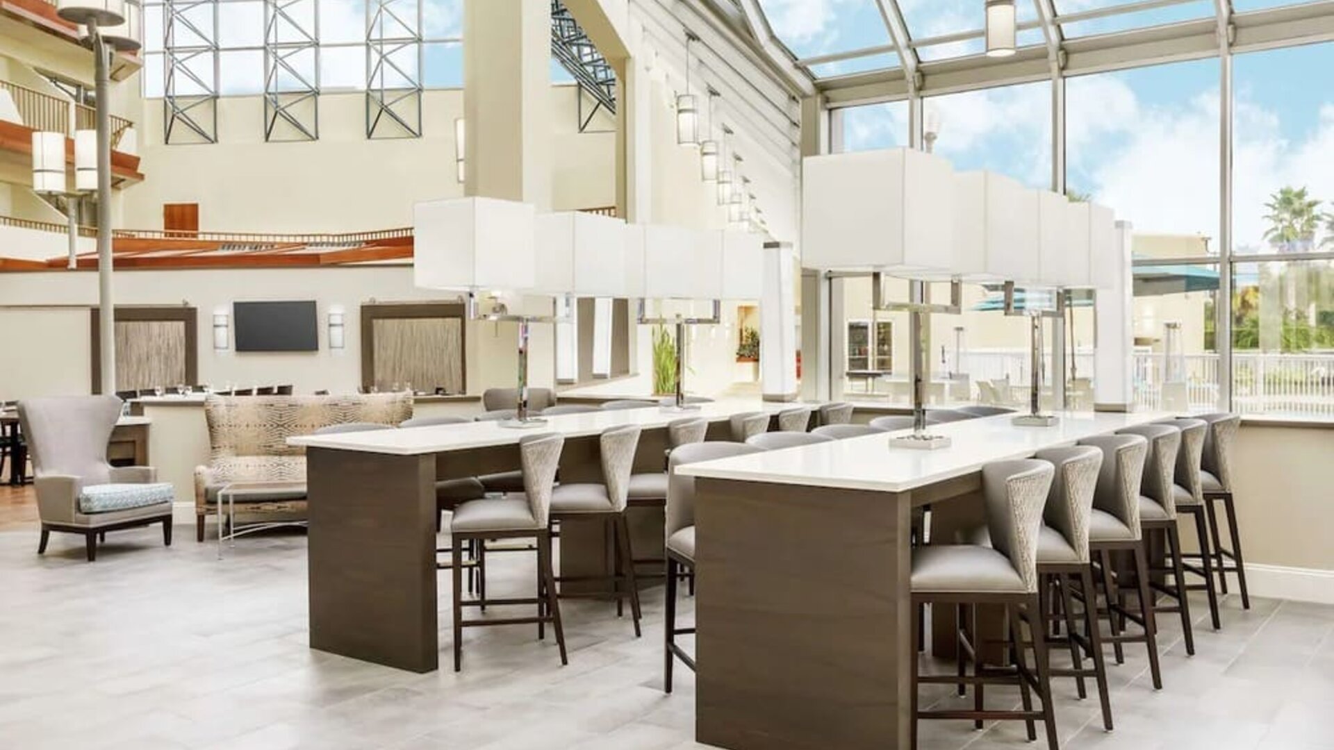 Bright atrium workspace with high-top communal tables, cushioned seating, floor-to-ceiling windows, and abundant natural light.